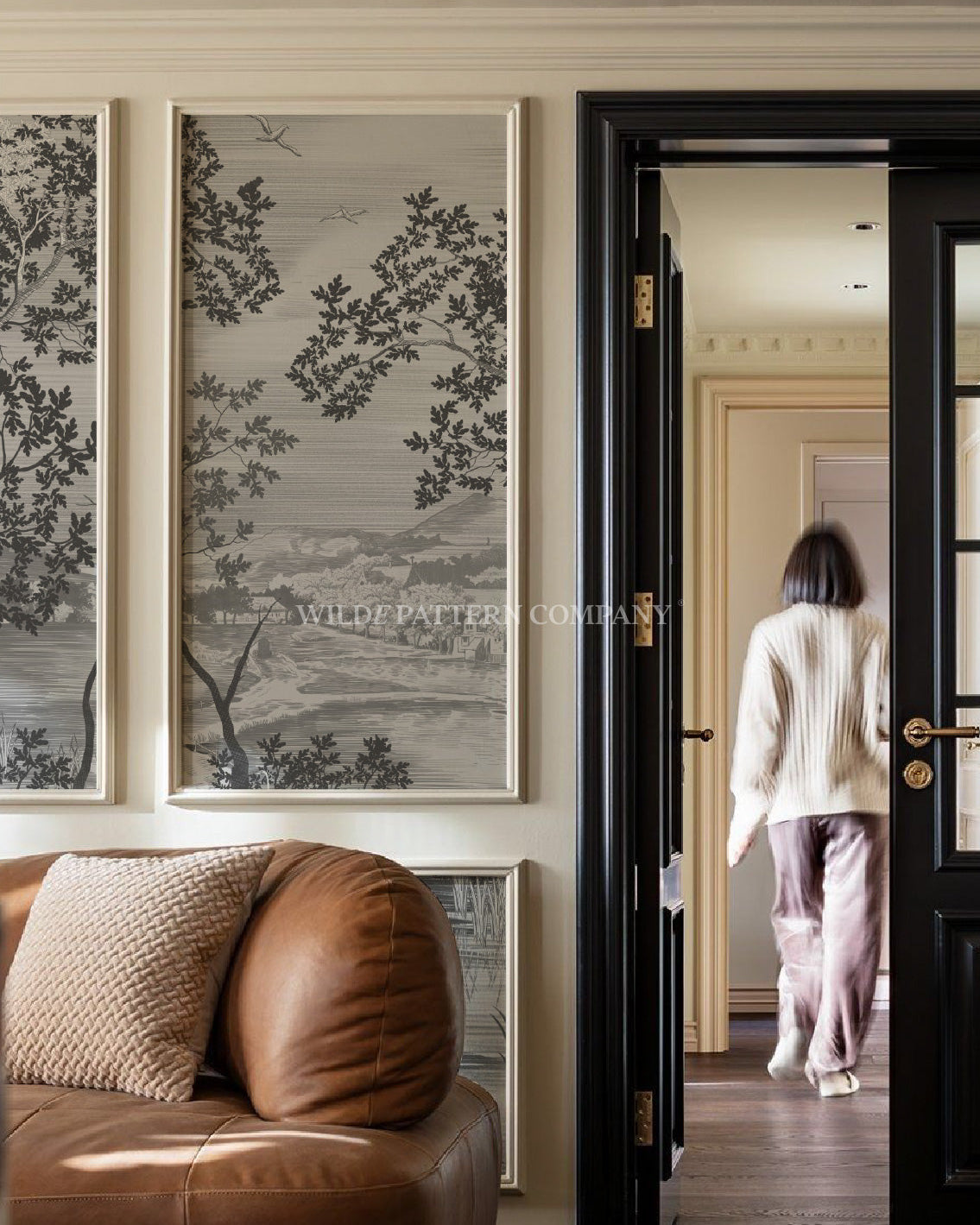 Living room with brown leather sofa, framed artwork on the wall, and a person walking through a doorway.
